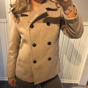 Beige skepparkavaj från H&M - Snygg beige skepparkavaj från H&M med dubbelknäppning och svarta knappar. Jackan har klassisk krage, raka fickor och är fodrad. Perfekt för dig som vill ha en stilren och tidlös look till höst och vinter.