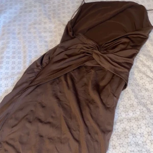 Brun axelbandslös omlottklänning - Snygg brun axelbandslös klänning med omlottdetalj framtill. Materialet har en lätt glans och känns mjukt mot huden. Perfekt för dig som gillar en stilren look med twist. Klänningen har en smickrande passform och knytdetalj vid midjan.