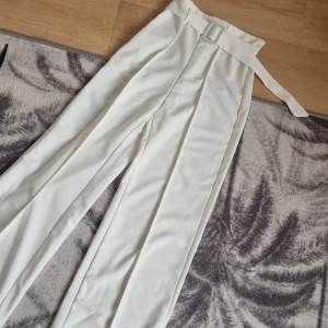 Vita kostymbyxor med bälte - Snygga vita kostymbyxor med hög midja och tillhörande bälte. Byxorna har raka ben och pressveck framtill för en clean look. Perfekta att styla med en crop top eller oversized skjorta för en trendig vibe. 