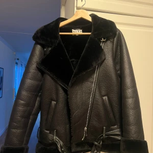 Svart pälsjacka/Pilotjacka från NA-KD  - Säljer en svart pälsjacka från NA-KD med bred krage, synlig dragkedja och bälte i midjan. Jackan har en oversized passform, långa ärmar och mjukt fuskpälsfoder. Perfekt för dig som vill ha en cool och varm look under hösten och vintern.