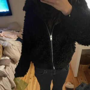 Svart teddyjacka med dragkedja - Mysig svart teddyjacka med fluffig yta och hel dragkedja framtill. Jackan har en avslappnad passform och långa ärmar, perfekt för att hålla värmen. Storlek L men passar mig som S, beroende på hur man vill att den ska sitta💕