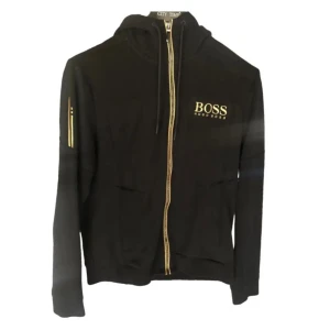 Svart hoodie från Hugo Boss med guld - Svart hoodie från Hugo Boss med guldfärgade detaljer och dragkedja. Tröjan har huva med snörning, guldlogga på bröstet och fickor framtill. Perfekt för dig som gillar stilrena och exklusiva plagg med en fräsch touch. PRIS KAN DISKUTERAS