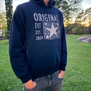 Mörkblå hoodie med stjärntryck - Snygg mörkblå hoodie från Original Fit med stort tryck framtill med stjärnor och texten 'Original Fit Est. 2024'. Tröjan har huva, magficka och långa ärmar. Perfekt för dig som gillar en avslappnad och cool stil.