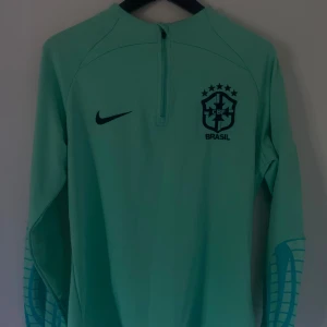 Fotbollströja - Snygg ljusgrön långärmad fotbollströja från Nike med Brasil-logga och svart swoosh på bröstet. Tröjan har halv dragkedja framtill och coolt mönster på ärmarna. Perfekt för dig som gillar landslagsstil och sportig look.