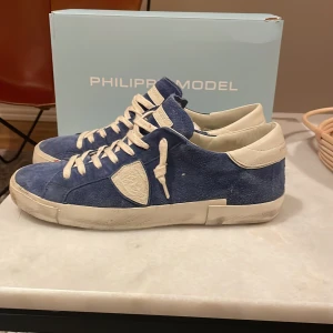 Blå Philippe Model sneakers mocka - ( Ny pris 3600kr) Snygga blå sneakers från Philippe Model i mocka med vita skinn-detaljer och vit sula. Skorna har klassisk snörning och en sköldlogga på sidan. Perfekta för dig som gillar stilrena och trendiga sneakers med premiumkänsla. Om de är något ni undrar tveka inte av att höra av er. ( Använd 1-2 gånger)