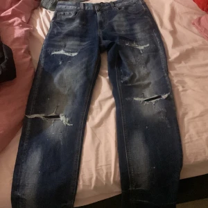 Blå distressed jeans Supply & Demand - Säljer ett par blå jeans från Supply & Demand med slitna detaljer och hål på benen. Jeansen har klassisk femficksdesign, normal passform och raka ben. Perfekt för dig som gillar en edgy streetstil. Materialet är klassisk denim.