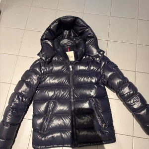 Mörkblå Moncler Maya pufferjacka med luva - Säljer en mörkblå pufferjacka från Moncler med glansig finish och huva. Jackan har dragkedja framtill, två snedställda fickor med dragkedja och Moncler-logga på ärmen. Insidan har en unik serietidningsetikett. Super varm och skön jacka som passar till allt! 💙köpt från barnavdelningen från Nk förra året helt ny och den är i den största storleken i barn så den passar som en vanlig S storlek! Visar självklart kvitto osv de är bara att skriva!🤗