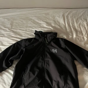 Svart vindjacka från Helly Hansen - Svart vindjacka från Helly Hansen med HH-logga på bröstet. Jackan har dragkedja framtill, två sidofickor med dragkedja och meshfoder på insidan. Perfekt för blåsiga dagar och enkel att matcha med olika outfits. Vill även tilllägga att storleken är 152 men jag som är över 165 har kunnat använda den utan att den är för liten. Nypris på jackan är 2000kr och har varit sparsamt använd                  / mvh Kasper 