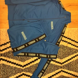 Blåa dress från Hoodrich - Snygga blåa mjukisbyxor från Hoodrich med elastisk midja och dragsko. Byxorna har svarta sidopaneler med Hoodrich-logga i vitt längs benen och tryckt logga på låret. 