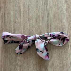 Blommigt rosa hårband med rosett - Snyggt hårband i mjukt rosa tyg med blommigt mönster i lila och grönt. Diademet har en stor rosett framtill som ger en gullig och trendig look. Perfekt för att piffa upp frisyren och ge lite extra färg till din outfit.