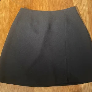 Svart kortkjol - Stilren svart kortkjol med rak passform och slits framtill. Kjolen har en dold dragkedja bak. Materialet är slätt och funkar till vad som helst💕