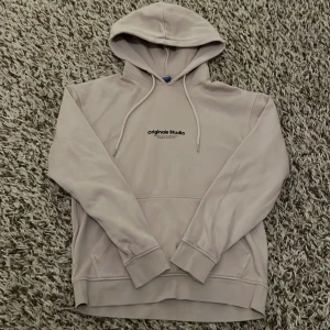 Beige hoodie från Jack & Jones - Säljer en stilren beige hoodie från Jack & Jones Originals med svart tryck 'originals Studio' på bröstet. Hoodien har huva med snörning, magficka och ribbade muddar. Perfekt för chill dagar och enkel att matcha med det mesta.