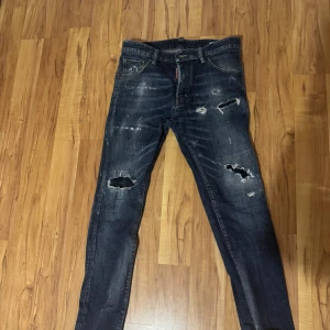 Blå slitna jeans från Dsquared2 - Säljer ett par blå jeans från Dsquared2 med coola slitningar och hål på knäna. Jeansen har klassisk femficksdesign, röd logotagg vid gylfen och en röd patch bak i midjan. De är raka i modellen och har en snygg tvättad look.