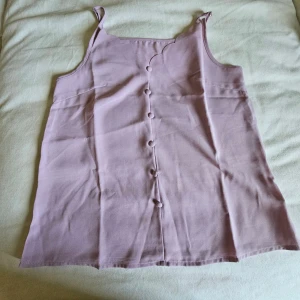 Ljuslila linnetopp med knappar - Säljer en ljuslila linnetopp med axelband och dekorativa knappar framtill. Toppen har en söt scallop-kant vid halsen och är tillverkad i mjukt bomullsmaterial. Perfekt för varma dagar och enkel att matcha med jeans eller kjol.