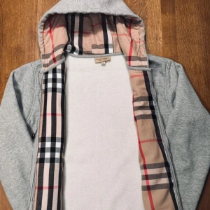 Grå hoodiejacka från Burberry - Grå hoodiejacka från Burberry med klassiskt rutigt foder i huvan och insidan. Jackan har dragkedja, snörning i huvan och två fickor framtill. Materialet är mjukt och bekvämt, perfekt för chill dagar. Den ikoniska Burberry-rutan ger en lyxig touch.