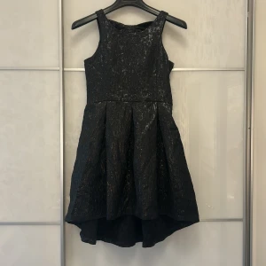 Svart glittrig kortklänning med rosett - Supersnygg svart kortklänning med glittrig yta och struktur. Klänningen har ärmlös design, markerad midja och voluminös kjol med lite längre bakstycke. Baksidan har en stor rosett och en v-ringad detalj, samt dragkedja i sidan för enkel påklädning.