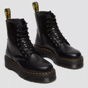 Svarta Dr. Martens boots i skinn - Klassiska svarta Dr. Martens boots i blankt skinn med gul söm runt sulan och grov platt sula. Skorna har snörning framtill, rund tå och den ikoniska gula logotaggen bak. Perfekta för dig som vill ha en edgy och tidlös look.