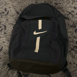 Svart ryggsäck från Nike - Säljer en marinblå ryggsäck från Nike med vit logga och detaljer på framsidan. Väskan har vadderade axelremmar och flera fack för smart förvaring. Perfekt för skola, träning eller vardag. Klassisk och sportig design.