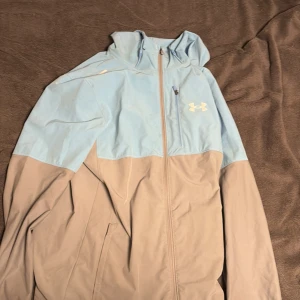 Under Armour vindjacka i blått och beige - Snygg vindjacka från Under Armour i ljusblått och beige. Jackan har huva, dragkedja framtill och praktiska fickor. Perfekt för dig som gillar sportig stil och vill ha något lätt och smidigt till vår eller höst.
