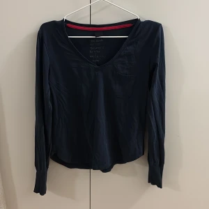 Mörkblå långärmad tröja med v-ringning - En stilren mörkblå långärmad tröja med v-ringad hals och liten bröstficka. Tröjan har en enkel design och en röd detalj på insidan av kragen. Perfekt för en chill och avslappnad look.