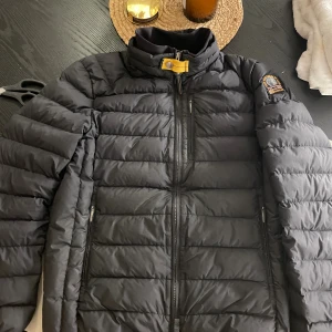 Svart dunjacka Parajumpers herr S - Säljer en svart dunjacka från Parajumpers i herrmodell, storlek L. Jackan är superlätt, har gul detalj vid kragen, dragkedjor på fickor och bröstficka samt klassisk Parajumpers-logga på ärmen. Perfekt för kyliga dagar och riktigt snygg passform. defekterna som finns. Syns på bild