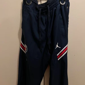 PSG x Jordan marinblå träningsbyxor - Marinblå träningsbyxor från PSG x Jordan med vita och röda ränder på benen, PSG-logga och Jumpman-logga. Byxorna har elastisk midja med snörning och dragkedjefickor. Perfekta för sport eller chill.