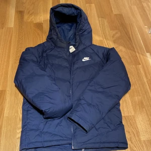 Marinblå pufferjacka från Nike 158-170 - Säljer en marinblå pufferjacka från Nike i storlek 158-170. Jackan har huva, dragkedja framtill och klassisk Nike-logga på bröstet. Perfekt för kalla dagar med sin vadderade design och sportiga look.