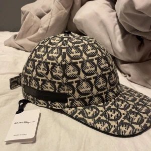 Salvatore Ferragamo mönstrad keps - Snygg keps från Salvatore Ferragamo i svart och beige med grafiskt mönster och justerbart spänne bak. Kepsen är tillverkad i bomull och har en läderdetalj med präglad logga. Perfekt accessoar för att lyfta din outfit.