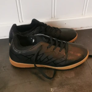 Svarta Umbro fotbollsskor med gummisula - Säljer ett par svarta fotbollsskor från Umbro med snygga grafiska detaljer på sidan och klassisk snörning. Skorna har en ljusbrun platt gummisula som ger bra grepp på inomhusplaner. Ovandelen är i syntetmaterial med en stilren look och rund tå. Skorna är bara änvända 2 gångef