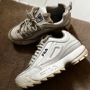 Vita chunky sneakers från FILA - Säljer ett par vita chunky sneakers från FILA med grov sula och snörning. Skorna har en cool retrodesign med FILA-logga på sidan och tungan, samt detaljer i rött och blått. Tillverkade i syntetmaterial och mesh för extra komfort och stil. 