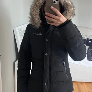 Svart pufferjacka Horse Legend - Svart pufferjacka från Horse Legend Wear med avtagbar huva och fuskpäls. Jackan har dragkedja, knappar och två fickor med dragkedja. Broderad logga på bröstet och patch på ärmen. Perfekt för kalla dagar.