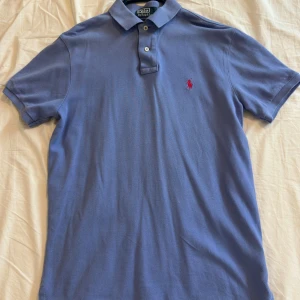 Blå pikétröja från Polo Ralph Lauren - Snygg blå pikétröja från Polo Ralph Lauren med klassisk krage och två knappar framtill. Tröjan har korta ärmar och den ikoniska röda Polo-loggan broderad på bröstet. Perfekt för en chill och stilren look.