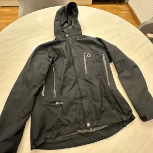  Haglöfs jacka - Svart dam vindjacka från Haglöfs i Gore-Tex-material med huva och flera dragkedjeförsedda fickor på bröstet. Jackan har en sportig look, justerbar nederkant och diskreta loggor. Perfekt för dig som gillar outdoor och vill ha en stilren jacka.