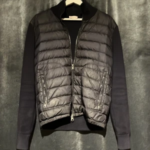 Svart pufferjacka från Moncler - Svart pufferjacka från Moncler med quiltad front och släta ärmar. Jackan har två dragkedjefickor, ribbade muddar och klassisk Moncler-logga på ärmen. Materialmix med polyester och bomull ger en sportig och clean look. Perfekt för kyliga dagar. Kvitto finns