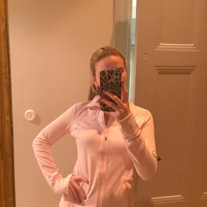 Ljusrosa sportig vindjacka - Säljer en ljusrosa vindjacka med dragkedja framtill och två fickor med dragkedja. Jackan har en sportig look, smal passform och är tillverkad i ett tunt, mjukt syntetmaterial. Perfekt för vår eller sommar när du vill ha något lätt och snyggt.