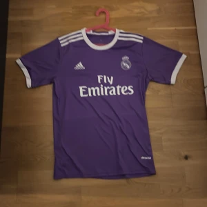 Lila Real Madrid fotbollströja Ronaldo - Säljer en lila Real Madrid fotbollströja från Adidas med Ronaldo 7 på ryggen. Tröjan har vita detaljer vid krage, ärmslut och axlar samt tryck med Fly Emirates på bröstet. Klassisk kortärmad modell i lätt och ventilerande material. Priset är ej hugget i sten 
