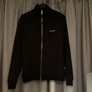 Svart track jacket från Les Deux - Snygg svart track jacket från Les Deux med silvrig dragkedja och diskret logga på bröstet. Jackan har hög krage, raka muddar och är tillverkad i ett mjukt polyestertyg. Perfekt för en clean och snygg stil. Nypris: 1200kr