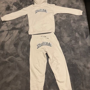 Beige hoodie från Hoodrich - Säljer en beige hoodie från Hoodrich med svart och vit logga framtill. Tröjan har huva med tryck, känguruficka och långa ärmar. Mjuk och skön bomullsmix, perfekt för chill eller streetwear. Matcha med tillhörande byxor för en komplett look.