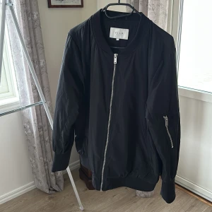 Svart bomberjacka från VILA - Svart bomberjacka från VILA med klassisk rund krage, silverfärgade dragkedjor framtill och på ärmen. Jackan har ribbade muddar vid ärmslut och nederkant, samt en stilren och enkel design. Perfekt för dig som gillar en clean och urban look.