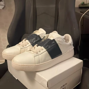 Valentino Garavani Open sneakers vit/blå - Säljer ett par Valentino Garavani Open sneakers i vitt skinn med bred marinblå rem över vristen. Klassisk rund tå, platt sula och vita snören. Märkeslogga på plösen. Skorna kommer med originalkartong, dustbag och äkthetskort.