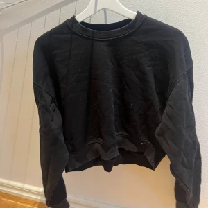 Svart croppad sweatshirt - En svart croppad sweatshirt med rund hals och långa ärmar. Tröjan har en avslappnad passform och ribbade muddar vid ärmslut och nederkant. Perfekt för en chill och stilren look.