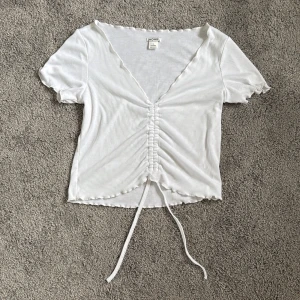 Vit ribbad topp med snörning från Monki - Superfin vit ribbad topp från Monki i storlek S. Modellen har korta ärmar, v-ringning och snygg snörning framtill som kan justeras för en croppad look. Toppens kanter har vågig finish för extra detalj. Perfekt till jeans eller kjol. Lite genomskinlig ska tilläggas, annars super fin att piffa upp med smycken. 