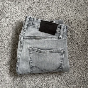 Gråa jeans från Jack & Jones (28/30) - Snygga ljusgrå jeans från Jack & Jones med klassisk femficksdesign och diskret broderad bakficka. Modellen är tapered med normal passform och midwaist. Jeansen är tillverkade i mjukt bomullsmaterial och har metallknappar och dragkedja framtill.