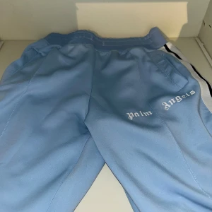 Palm Angels ljusblå mjukisbyxor - Snygga ljusblå mjukisbyxor från Palm Angels med vit och svart sidoremsa. Byxorna har elastisk midja, fickor och logotypen tryckt på framsidan. Perfekt för chill eller streetwear. Materialet är mjukt och skönt, troligtvis polyester.