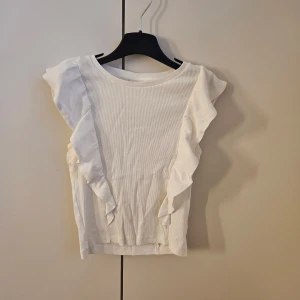 Vit ribbad topp med volang från Kappahl - Säljer en vit ribbad topp från Kappahl i storlek 158/164. Toppen har snygga volangdetaljer längs axlarna och är ärmlös. Materialet är mjuk bomull och modellen är normal passning. Perfekt för dig som gillar stilrena och feminina detaljer.