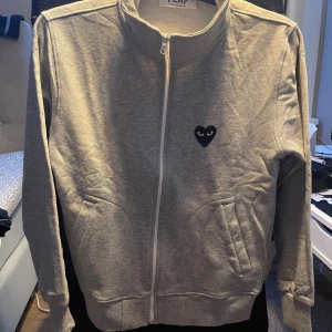 Grå zip up CDG grå - Grå ziptröja från cdg med hjärta på bröstet och baksidan. Tröjan har hög krage, långa ärmar och två fickor framtill.