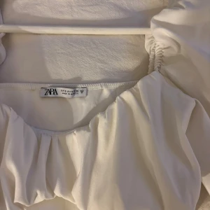 Vit croppad blus med puffärm från Zara - Supersnygg vit blus från Zara med croppad passform och puffiga långärmar. Toppen har en fyrkantig urringning och rynkade detaljer framtill som ger en cool och trendig vibe. Perfekt att styla med höga jeans eller kjol för en chic look. Finns en liten fläck som ni ser på sista bilden, går säkert bort i tvätten men lovar inget.