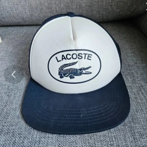 Vit och marinblå Lacoste keps - Snygg keps från Lacoste i vit och marinblå färg med klassisk krokodillogo framtill. Framsidan är vit med tryckt logga, skärmen och mesh-panelerna är marinblå. Perfekt för dig som vill ha en sportig och stilren look.
