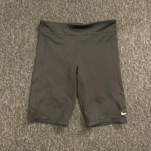 Svarta Dri-Fit träningsshorts Nike S - Svarta, tighta träningsshorts från Nike i Dri-Fit material. De har hög midja och en stilren design med Nike-loggan i silver på benet. Perfekta för gymmet eller löpning, med stretchigt syntetmaterial som andas och sitter skönt. Det står att shortsen är i XL i barnstorlek så skulle säga att de passar någon som har S.