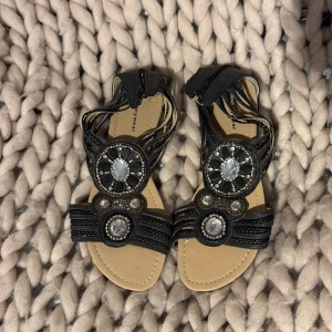 Svarta sandaler med pärldetaljer Mywear - Svarta sandaler från Mywear i storlek 41 med platt sula och rund tå. Dekorerade med stora och små pärlor och glittriga stenar i cirkelmönster framtill. Remmarna är tunna och korsar över foten för en snygg look. Perfekta för sommarens alla tillfällen.